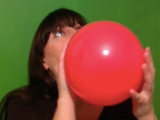 Eine Person hält einen roten Luftballon vor einem leuchtend grünen Hintergrund und bläst ihn auf.