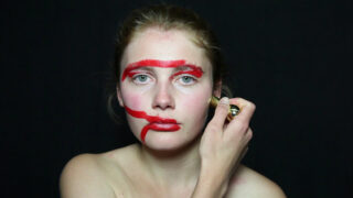 Eine Person trägt roten Lippenstift in dicken, ungleichmäßigen Linien quer über ihr Gesicht auf, mit einem schwarzen Hintergrund dahinter.