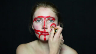 Eine Person trägt vor einem schlichten schwarzen Hintergrund ein kräftiges rotes Make-up in geometrischen Mustern auf ihr Gesicht auf.