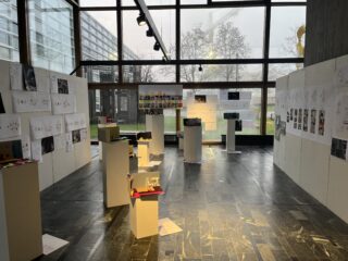 Raum mit großen Fenstern, in dem Architekturmodelle auf Sockeln und Konstruktionszeichnungen an den Wänden ausgestellt sind, mit natürlichem Lichteinfall von außen.