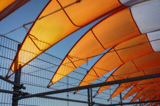 Orangefarbene und weiße Stoffplanen sind über einen Metallrahmen gespannt, so dass geschwungene Formen entstehen und der blaue Himmel durch einen Drahtzaun im Hintergrund zu sehen ist.