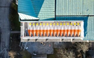 Luftaufnahme von elf kleinen Segelbooten mit gelben und orangefarbenen Abdeckungen, die in einer Reihe auf einem Dach in der Nähe eines grünen Gebäudes mit Metalldächern angeordnet sind.