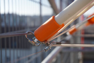 Nahaufnahme einer orangefarbenen und weißen Stange, die mit einem Metallhaken und einer Klemme an Metallkabeln befestigt ist, vor einem unscharfen Hintergrund im Freien mit einem Zaun.