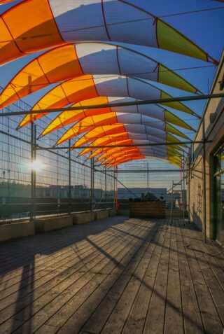 Auf der Dachterrasse mit Holzboden sind gelbe und weiße Sonnensegel angebracht, die in der späten Nachmittagssonne gemusterte Schatten werfen.