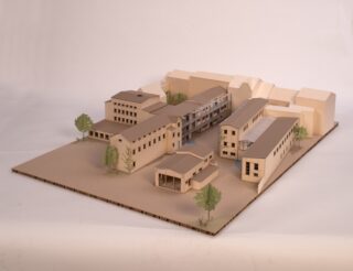 Ein maßstabsgetreues Architekturmodell zeigt mehrere beigefarbene Gebäude, die um einen zentralen Innenhof angeordnet sind, mit spärlichen Bäumen und einfachen Landschaftselementen.