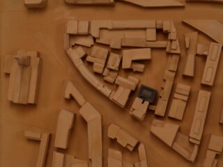 Ein braunes Architekturmodell eines Stadtblocks mit verschiedenen rechteckigen und unregelmäßigen Gebäuden; ein Gebäude in der Nähe der Mitte ist schwarz hervorgehoben.