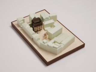 Architektonisches Modell im Maßstab 1:1, das eine Gruppe hellgrüner Gebäude mit einem zentralen braunen Gebäude auf einem rechteckigen Holzsockel vor einem weißen Hintergrund zeigt.