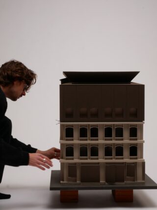 Eine Person justiert ein detailliertes architektonisches Modell eines mehrstöckigen Gebäudes, das auf einer ebenen Fläche steht, die von zwei Holzklötzen getragen wird.