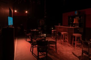 Schummrig beleuchtete Bar mit leeren Stühlen und Hockern, runden Tischen, Kerzen und einem Fernsehbildschirm an der linken Wand. Auf dem Bartresen stehen Regale und eine Lampe. Die Szene scheint unbewohnt zu sein.