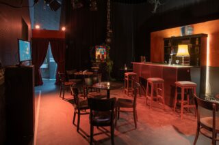 Schummrig beleuchtete Bar mit Tresen, Hockern, mehreren Tischen und Stühlen, einer Lampe auf dem Tresen, Regalen mit Flaschen und einem Vorhang, der in einen anderen Raum führt.