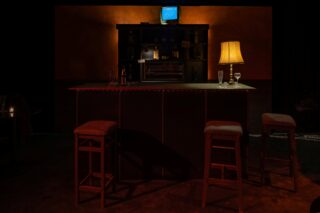 Eine schwach beleuchtete Bar mit drei leeren Hockern, einem Bartresen mit Gläsern und einer Lampe, Regalen mit Flaschen und einem kleinen Fernseher im Hintergrund.