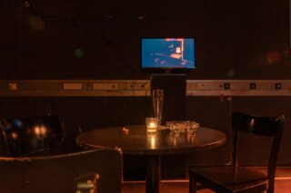 Ein schwach beleuchteter Raum mit einem runden Tisch, zwei Stühlen, einem leeren Glas, einer brennenden Kerze, einem Aschenbecher und einem kleinen Fernseher, der im Hintergrund eine dunkle Szene zeigt.