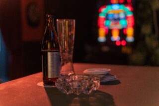 Eine Bierflasche, ein leeres Glas, ein Aschenbecher und ein Stapel Untersetzer stehen auf einem Bartresen mit einem verschwommenen bunten Licht im Hintergrund.