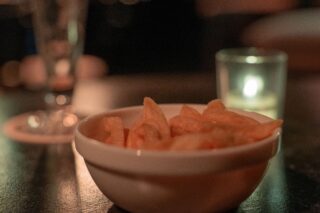 Auf einem dunklen Tisch steht eine weiße Schale mit Kartoffelchips, daneben eine brennende Kerze und ein leeres Glas.