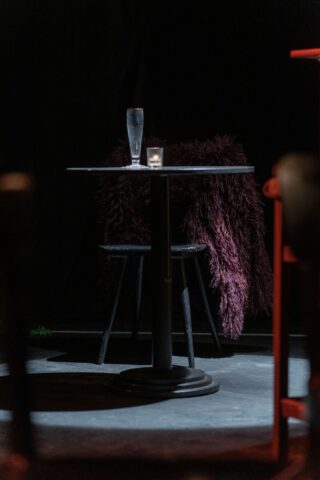 Ein kleiner runder Tisch mit einem Glas Wasser, einer brennenden Kerze und einem Stuhl, der mit einem dunklen, pelzigen Stoff bezogen ist, alles vor einem dunklen Hintergrund.