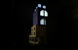 Ein kleines Architekturmodell eines Gebäudes mit beleuchteten oberen Stockwerken und großen Bogenfenstern vor einem dunklen Hintergrund.