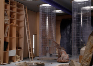 Das moderne Interieur besteht aus hohen transparenten Zylindern, einer Holzleiter, Regalen mit verschiedenen Gegenständen und der Skulptur einer sitzenden Figur, die von Felsen umgeben ist.