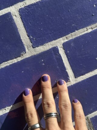 Eine Hand mit silbernen Ringen und lila lackierten Nägeln berührt eine lila Backsteinmauer mit weißem Mörtel.