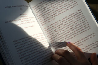 Eine Hand hält ein Buch mit englischem und deutschem Text aufgeschlagen, das teilweise von Sonnenlicht und Schatten beleuchtet wird.