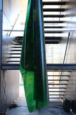 Ein großes grünes, durchscheinendes Blatt hängt vertikal in der Mitte einer modernen Metalltreppe mit offenen Setzstufen und einer industriellen Umgebung.