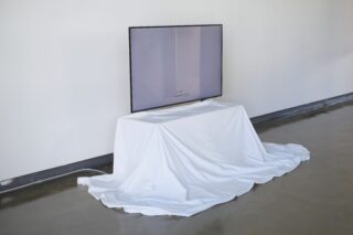 Ein Flachbildfernseher steht auf einem mit einem weißen Laken bedeckten Tisch an einer schlichten Wand, wobei der Bildschirm leer ist.