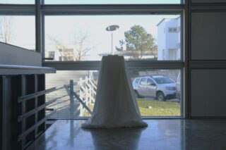 Ein mit einem weißen Tuch bedeckter Sockel steht vor einem großen Fenster, vor dem bei Tageslicht ein Auto und Häuser zu sehen sind.