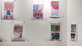 Sechs farbenfrohe, handgezeichnete Poster mit abstrakten Mustern und handgeschriebenem Text sind auf einer weißen Galeriewand ausgestellt, jedes mit kleinen Notizen darunter an der Wand befestigt.