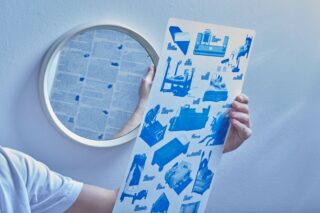 Eine Person hält ein Blatt mit blauen Abbildungen verschiedener Gegenstände neben einen runden Spiegel, der eine Seite mit gedrucktem Text auf einer weißen Wand reflektiert.