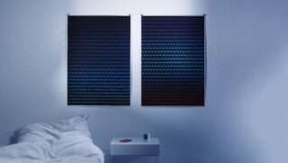 Ein ordentlich gemachtes Bett neben einem kleinen weißen Nachttisch mit einem roten Buch und einem Wecker, an einer Wand mit zwei geschlossenen Jalousien in schummrigem blauem Licht.