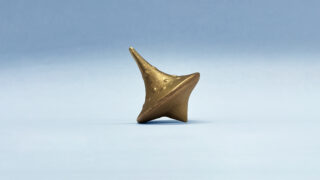 Ein bronzener Kreisel balanciert aufrecht auf einer ebenen Fläche vor einem schlichten hellblauen Hintergrund.