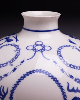 Nahaufnahme des oberen Teils einer weißen Porzellanvase, die mit komplizierten blauen Mustern, darunter Schleifen, Ketten und abstrakte Formen, vor einem dunklen Hintergrund verziert ist.