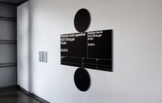 An der Wand montierte schwarz-weiße Schilder zeigen Daten für fertiges Motorenbenzin und destilliertes Heizöl an, einschließlich Preise und prozentuale Veränderungen, mit kreisförmigen Akzenten über und unter den Schildern.