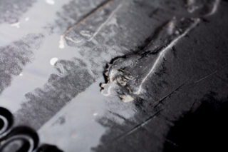 Nahaufnahme einer schwarzen Oberfläche mit Kratzern, Abnutzungsspuren und glänzenden Flecken einer klaren Flüssigkeit oder eines Rückstandes.