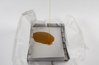 Eine dicke bernsteinfarbene Flüssigkeit wird auf einen Stapel von Dokumenten in einem Ordner gegossen, der teilweise mit zerknülltem Wachspapier bedeckt ist.