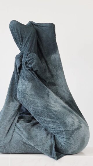 Eine große, dunkelgraue Decke wird zu einer hohen, abstrakten Form vor einem schlichten weißen Hintergrund gewickelt und gebunden.