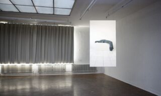 In einem minimalistischen Galerieraum mit grauen Vorhängen und braunem Boden hängt eine Fotografie. Das Foto zeigt eine in dunklen Stoff gehüllte Person, die mit dem Gesicht nach unten auf einem weißen Sockel liegt.