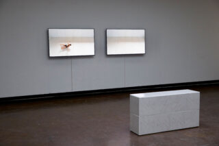 Eine minimalistische Galerie mit zwei Bildschirmen an einer weißen Wand zeigt gespiegelte Bilder einer Tänzerin, vor denen eine weiße rechteckige Bank auf einem dunklen Boden steht.