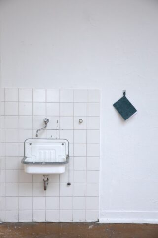 Ein kleines weißes Waschbecken mit einem Wasserhahn und einer Kette ist an einer gefliesten Wand angebracht; rechts davon hängt ein dunkelgrünes Reinigungskissen an einer weißen Wand.