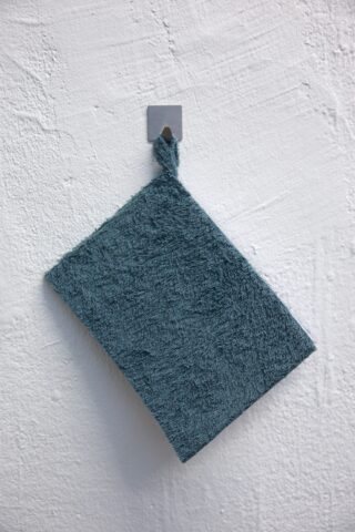 Ein blauer strukturierter Waschlappen mit einer Aufhängeschlaufe hängt an einem kleinen Metallhaken, der an einer weißen strukturierten Wand angebracht ist.