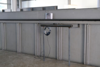 Ein schwarzer Kopfhörer hängt an einer Metallstange, die an einer grauen Trennwand in einem Raum im Industriestil befestigt ist.