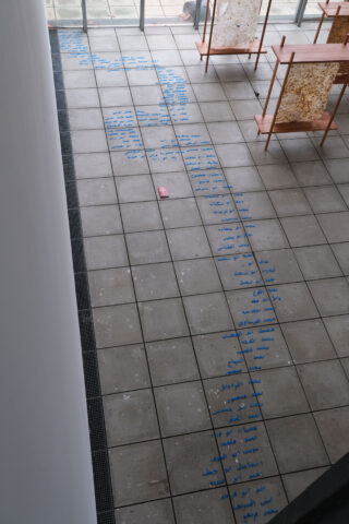 Blick von oben auf einen gefliesten Boden mit handgeschriebener blauer Schrift, die einen gewundenen Weg bildet, der zwischen hölzernen Ausstellungstischen in der Nähe großer Fenster verläuft; auf dem Boden liegt ein rosa Radiergummi.