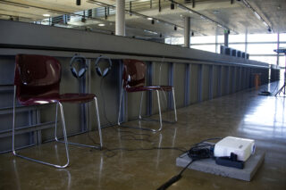 Zwei kastanienbraune Stühle mit Kopfhörern an den Rückenlehnen stehen nebeneinander in einem großen, modernen Raum. Vor ihnen steht ein Projektor auf dem Boden, dessen Kabel über den polierten Boden verlaufen.