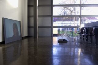 In einem Raum mit großen Fenstern, polierten Böden und mehreren Stühlen an einem Tresen wird das Bild einer Frau auf eine an die Wand gelehnte Tafel projiziert.
