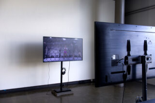 Ein Flachbildfernseher auf einem Ständer zeigt ein Sportereignis, darunter hängen Kopfhörer. Der Fernseher steht in einem kargen Raum vor einer Wand.