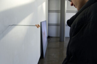 Eine Person in einer dunklen Jacke beobachtet eine Kunstinstallation mit Drähten, die an einem kleinen Objekt an einer weißen Wand befestigt sind, während im Hintergrund ein Bildschirm an die Wand gelehnt ist.