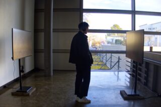 Eine Person mit Kopfhörern steht zwischen zwei großen Bildschirmen in einem Raum mit einem großen Fenster, das einen Blick auf einen Außenbereich bietet.