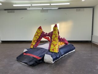 Eine aufblasbare Skulptur mit roten und gelben Bögen auf einem schwarz-weißen Sockel steht in einem leeren, gut beleuchteten Raum mit weißen Wänden und einem dunklen Boden.