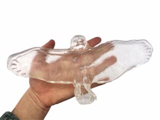 Eine Hand, die eine klare, glasartige Skulptur in Form eines Vogels mit ausgebreiteten Flügeln hält, vor einem weißen Hintergrund.