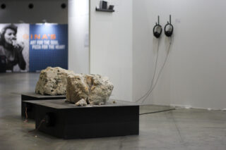 In einer Kunstgalerie sind zwei große Felsen auf schwarzen Podesten ausgestellt. An der angrenzenden weißen Wand sind Kopfhörer angebracht, und im Hintergrund ist ein verschwommenes Plakat zu sehen.