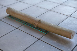 Ein zylindrischer Holzklotz wird auf einen gefliesten Boden gelegt und liegt der Länge nach auf einem rechteckigen grünen Lineal oder Messbrett.